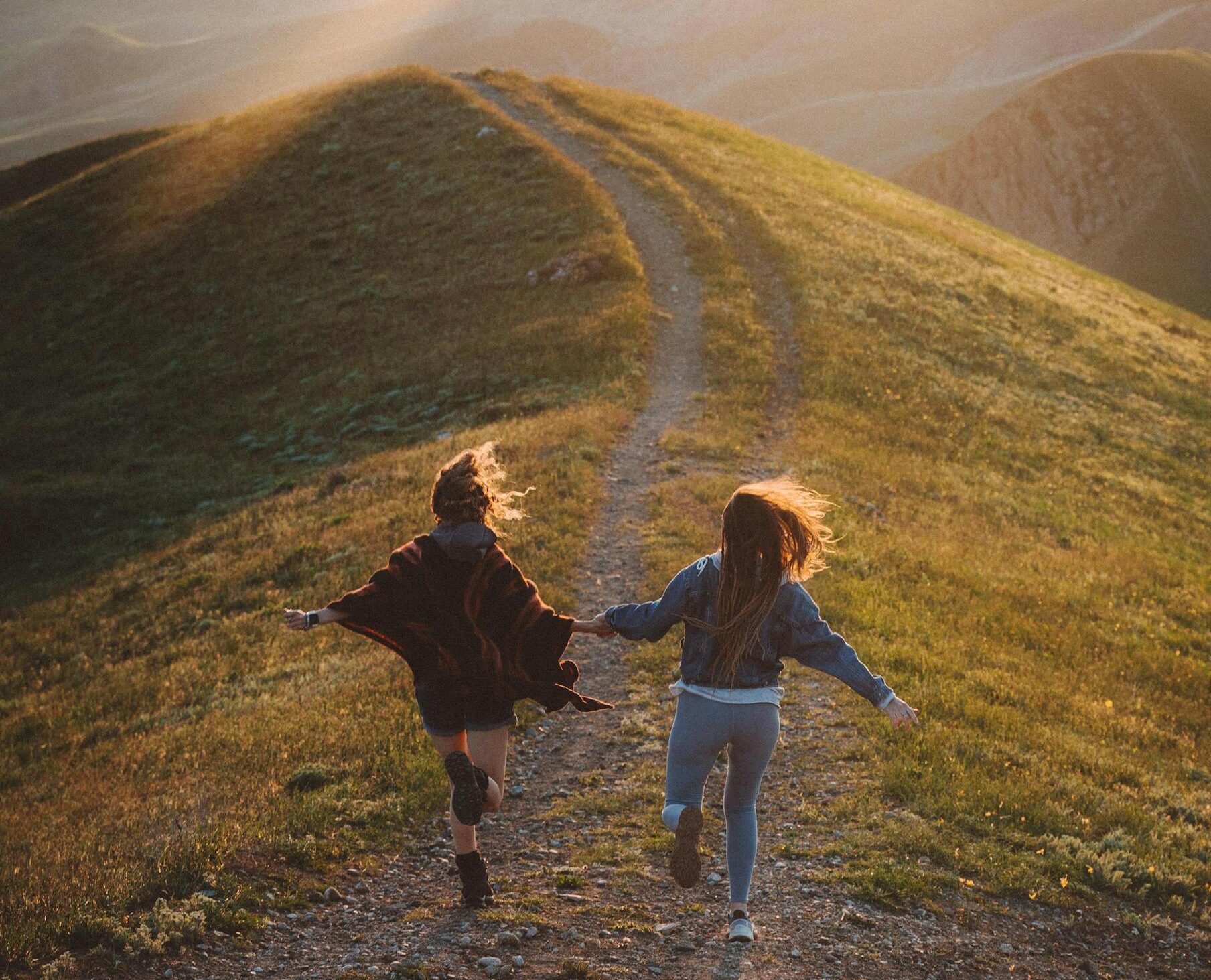 Two women running joyfully together on a scenic mountain trail during sunset.