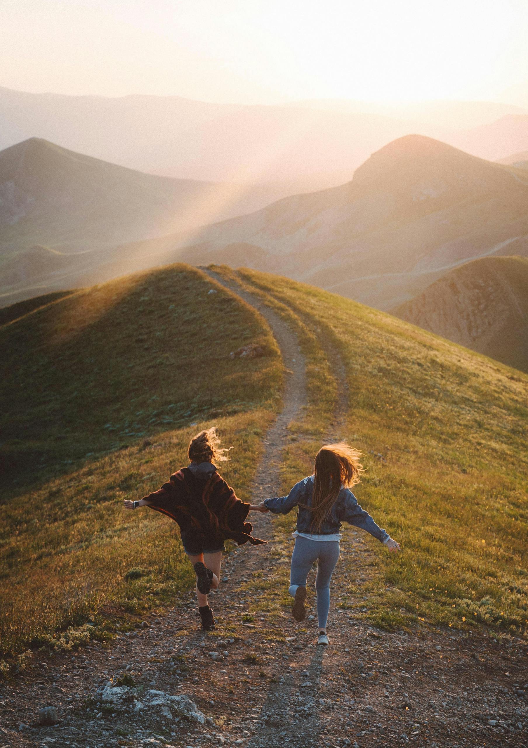 Two women running joyfully together on a scenic mountain trail during sunset.