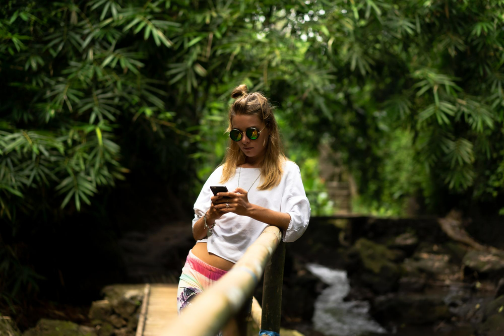 A woman stands in the nature and writes something on her phone, she wears a sunglasses and surrounded by the nature.