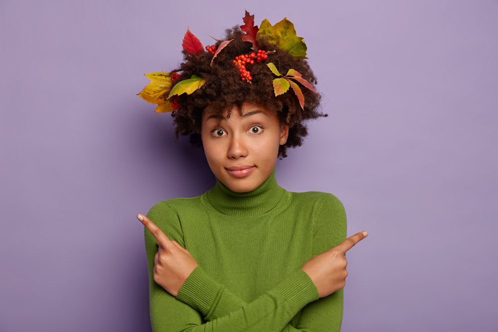 Illustration for the Frequently Aked Questions page. A black woman standing front of us in a green rolled neck pullover. Her hair is decorated with autumn leaves and her armes crossed in fron of her and pointing up with her pointing fingers.